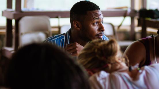 Black adult man sitting in a classroom-like environment.