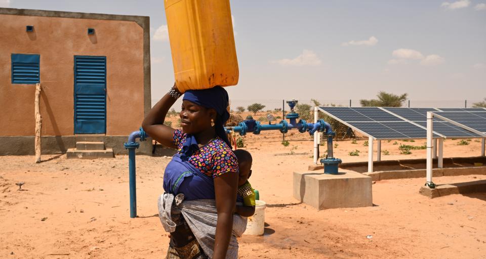 A woman in Africa, carrying a baby on her back, and holding a yellow water container on her head